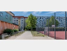 Gehweg zwischen einem rosa-orangefarbenen Gebäude links und einem eingezäunten Sportplatz mit rotem Bodenbelag rechts, im Hintergrund ein mehrstöckiges Wohngebäude mit bemalter Fassade, umgeben von Bäumen und Grünflächen. Im Vordergrund befinden sich Fahrradständer und eine Sitzbank.