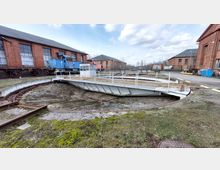 Historischer Lokschuppen mit Drehscheibe im Vordergrund, umgeben von roten Backsteingebäuden. Im Hintergrund sind ein blauer und ein orangefarbener Güterzug zu sehen.