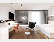 Modernes Hotelzimmer mit minimalistischem Design, ausgestattet mit einem großen Bett, einem schwebenden weißen Schreibtisch mit orangefarbenem Stuhl und einem an der Wand montierten Fernseher. Der Raum verfügt über Holzboden, eine Sitznische mit Betonwand und Spiegel sowie große Fenster mit grauen Vorhängen, die viel Tageslicht hereinlassen.
