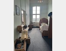 Schmaler Raum mit historischem Interieur, darunter ein Holzbett, ein alter Nähtisch mit Nähmaschine und eingelegte Textilien. Ein großes, bogenförmiges Fenster lässt Tageslicht herein; im Vordergrund steht ein Korb mit einer Puppe.