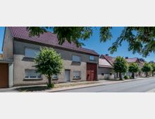 Straßenszene mit einer Reihe von Wohnhäusern mit roten Ziegeldächern und verputzten Fassaden. Vor den Gebäuden stehen kleine Bäume entlang eines Bürgersteigs, daneben eine asphaltierte Straße unter einem hellblauen Himmel.