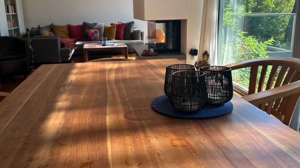 Heller Wohn- und Essbereich mit großem Holztisch im Vordergrund, schwarzem Kerzenhalter-Set und Pendelleuchten an der Decke. Im Hintergrund stehen Sofa und Couchtisch neben einem modernen Kamin; rechts führen bodentiefe Fenster mit Vorhängen zu einem grünen Garten bzw. Balkon.