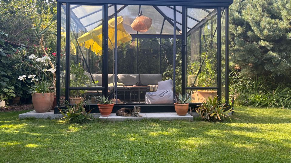 Schwarzes, verglastes Gartenhaus mit Satteldach in einem grünen Garten, umgeben von Bäumen und Sträuchern. Innen sind eine L-förmige Sitzgruppe und eine Hängelampe zu sehen, davor stehen mehrere Terrakotta-Blumentöpfe auf einer niedrigen Steinplatte.