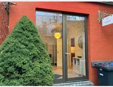 Rote Gebäudefassade mit großer Glastür, die einen Einblick in einen Innenraum mit heller Einrichtung und einer hängenden, gelben Lampe gibt. Vor dem Gebäude steht ein dichter, kegelförmiger Strauch; rechts davon hängt ein Schild mit der Aufschrift „Osteopathie Bianca Lachte“.