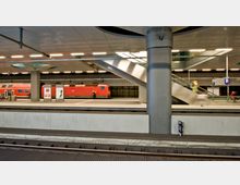 Ein moderner Bahnhof mit mehreren Ebenen, breiten Bahnsteigen und einem roten Regionalzug auf einem der Gleise. Eine Rolltreppe verbindet die Ebenen, und die Decke ist mit einer Metallverkleidung und Beleuchtung ausgestattet.