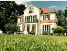 Zweistöckiges, weißes Wohnhaus mit rotem Ziegeldach, grünen Fensterläden und einem Balkon. Das Haus steht in einem Garten mit gepflegtem Rasen, Bäumen, Sträuchern und Gartenmöbeln.