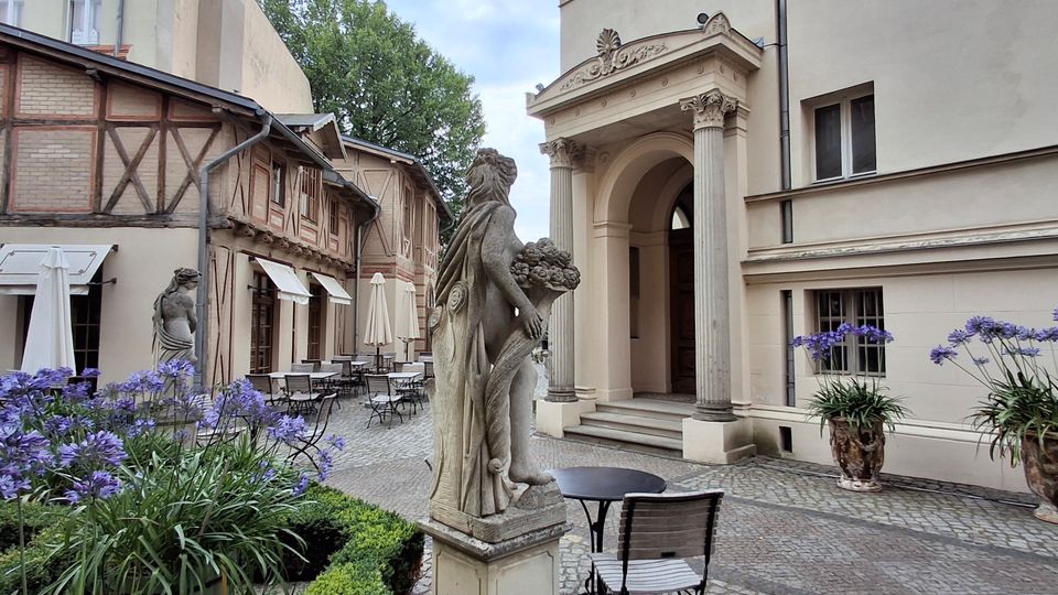 Innenhof mit Kopfsteinpflaster und Cafébestuhlung zwischen einem Fachwerkgebäude und einem klassizistischen Eingang mit Säulen und Rundbogen. Im Vordergrund stehen eine Steinfigur und blühende Pflanzen in Beeten und großen Töpfen.