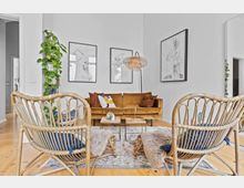 Wohnzimmer mit hellen Wänden, einer senfgelben Couch, zwei Rattan-Sesseln mit Fellauflagen und einem gemusterten Teppich auf einem Holzboden. An der Wand hängen vier abstrakte Schwarz-Weiß-Bilder, und links steht eine grüne Kletterpflanze.