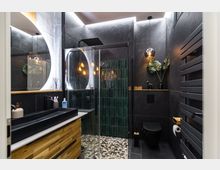 Modernes Badezimmer mit dunklen Wänden und einem großen, beleuchteten runden Spiegel. Die Dusche verfügt über grüne Fliesen, eine Glastür und einen Regenwaldduschkopf; daneben befindet sich ein schwarzes WC. Holz- und schwarze Elemente dominieren das Design, ergänzt durch dekorative Pflanzen und warmes Licht.