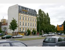 Straßenansicht eines vierstöckigen Gebäudes mit gelber Fassade und weiß gerahmten Fenstern, beschriftet mit "East-Side-Hotel". Im Vordergrund parken Autos, daneben steht ein Verkehrsschild mit Wegweisern, und im Hintergrund ist eine Baumreihe sichtbar.