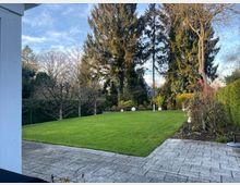 Ein gepflegter Garten mit einer Rasenfläche, umgeben von hohen Bäumen, Hecken und Sträuchern. Im Vordergrund ist eine gepflasterte Terrasse zu sehen, und vereinzelt sind Dekokugeln im Garten verteilt.