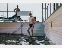 Ein Mann steht im knietiefen Wasser eines Pools und geht eine Treppe hinauf, während im Hintergrund ein zweiter Mann in Badebekleidung neben Liegestühlen steht. Die Szenerie zeigt einen modernen Außenbereich mit Holzvertäfelung und Metallgeländern.