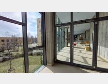 Ein heller Flur mit großen Glasfenstern und einer Glas-Doppeltür, die den Innen- und Außenbereich trennt. Draußen ist eine Grünfläche mit Bäumen und modernen Gebäuden im Hintergrund zu sehen, während drinnen gestapelte Stühle und Holzkisten stehen.