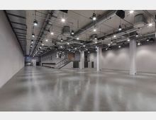 Großer, moderner, leerer Veranstaltungsraum mit einer glatten Betonbodenfläche, hohen weißen Wänden und sichtbaren Lüftungsrohren sowie mehreren Hängeleuchten an der Decke. Rechts befinden sich tragende Säulen, und links führt eine Treppe zu einer leicht erhöhten Plattform mit mehreren geschlossenen Türen.