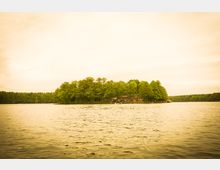 Eine kleine, bewaldete Insel auf einem ruhigen See, mit einem Gebäude am Ufer, das teilweise von Bäumen verdeckt ist; am Wasser befinden sich Stege und ein kleines Boot. Der Himmel ist bewölkt und das Licht wirkt gedämpft.