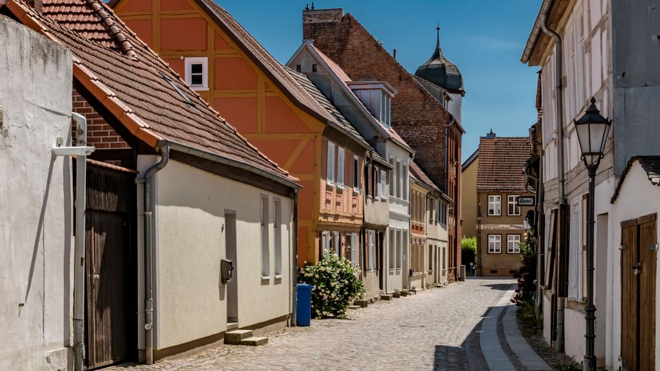 Schmale, gepflasterte Altstadtgasse mit dicht stehenden Häusern, teils Fachwerk- und teils verputzten Fassaden sowie roten Ziegeldächern. Die Straße verläuft leicht gebogen, mit Laterne am Rand und einem turmartigen Gebäude im Hintergrund.