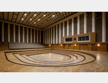 Ein leerer Konzert- oder Veranstaltungsraum mit hölzernem Boden, ansteigenden Sitzreihen im Hintergrund und einer Bühne in Halbrundform. Die Wände sind mit weißen Säulen und vertikalem Holz verkleidet, mit symmetrischen, gitterförmigen Fensterelementen und einer Uhr an der rechten Wand.