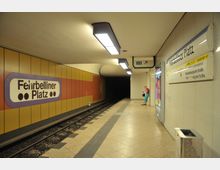 U-Bahn-Station Fehrbelliner Platz in Berlin, mit gelb-roten Wandpaneelen, einem Stationsschild und einer Person, die auf dem Bahnsteig steht. Im Hintergrund sind U-Bahn-Gleise und ein dunkel wirkender Tunnel sichtbar; eine Leuchtreklame und Wegweiser zur Linie U3 befinden sich rechts.
