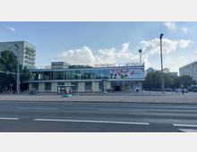 Straßenansicht eines modernen Gebäudes mit der Aufschrift „Moskau“, das als Restaurant und Veranstaltungsort dient. Das Gebäude hat eine Glasfassade im oberen Stockwerk, eine Werbetafel mit bunten Grafiken und ist von einer breiten, gepflasterten Fläche umgeben, auf der sich Fahrräder, Sitzbänke und einige Fußgänger befinden.