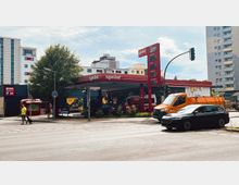 Straßenszene mit einer Tankstelle der Marke „Sprint“ im Vordergrund, umgeben von mehrspurigen Straßen. Im Hintergrund sind Wohn- und Geschäftsgebäude zu sehen, daneben ein Supermarkt mit „REWE“-Beschilderung. Ein Baum, parkende Fahrzeuge und Fußgänger prägen das Stadtbild.