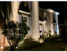 Nachtszene eines zweistöckigen Gebäudes mit heller Fassade, weißen Fensterläden und beleuchteter Eingangstür. Vor dem Haus befinden sich Sträucher und kleine Bäume in Töpfen, die durch Bodenlichter angestrahlt werden.