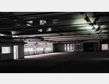 Große, leere Industriehalle mit sichtbaren Deckenstreben und Belüftungsrohren. Mehrere Fenster an den Seitenwänden lassen diffuses Licht in den ansonsten dunklen Raum.