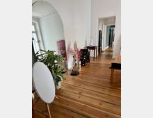 Ein stilvoll eingerichteter Innenraum mit Holzdielenboden, einem großen, halbkreisförmigen Spiegel, Zimmerpflanzen und dekorativen Elementen wie Trockenblumen. Im Hintergrund führen weiße Türrahmen zu weiteren Räumen, und an den Wänden ist schlichte weiße Gestaltung mit Stuckdetails sichtbar.