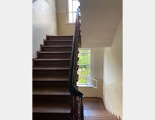 Treppenhaus in einem älteren Gebäude mit braunen Holzstufen und einem Holzgeländer. Ein großes Fenster mit geöffnetem Rahmen bietet Blick auf einen grünen Garten.