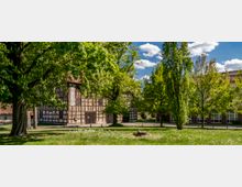 Fachwerkhaus in einem grünen Park mit großen Bäumen und Rasenflächen, daneben ein modernes Gebäude mit roten Dachziegeln und großen Fenstern. Der Himmel ist blau mit vereinzelten Wolken.
