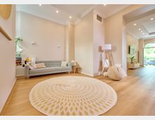 Wohnzimmer mit hellbeigen Wänden, Holzfußboden und moderner Einrichtung. Im Vordergrund ein runder Teppich mit geometrischem Muster, eine graue Couch an der Wand, eine Stehlampe mit Holzstativ und große Fenster, die Tageslicht hereinlassen.