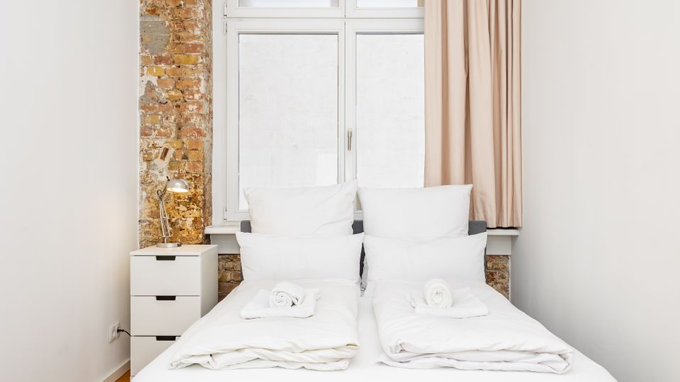 Schlichtes Schlafzimmer mit zwei zusammengestellten Einzelbetten in weißer Bettwäsche vor einem großen Fenster mit Vorhang. Links stehen ein kleiner weißer Nachttisch mit Lampe und eine freigelegte Backsteinwand als Akzent neben weißen Wänden.