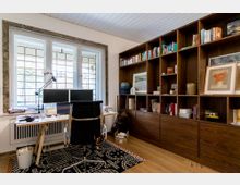 Ein Home-Office mit einem großen Fenster mit mehrfach geteiltem Glas im Hintergrund, davor ein Schreibtisch mit zwei Monitoren und einem schwarzen Bürostuhl. Rechts an der Wand steht ein deckenhohes Regal aus dunklem Holz, gefüllt mit Büchern, Bildern und Dekorationsgegenständen.