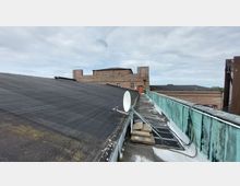 Flachdach eines Gebäudes mit schwarzer Dachabdeckung und darauf montierter Satellitenschüssel. Im Hintergrund ist ein mehrstöckiges, aus Backstein gebautes Gebäude mit Fenstern und flachen Dächern zu sehen, daneben eine grüne Metallverkleidung längs des Dachs.