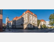Straßenszene mit einem großen historischen Gebäude aus hellem Stein und rotem Walmdach, flankiert von Bäumen und modernen Gebäuden im Hintergrund. Links im Bild ragt ein Kirchturm mit spitzem Dach hervor, das in den blauen Himmel ragt.