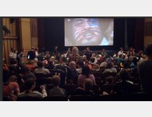 Ein Kinosaal mit zahlreichen sitzenden Personen, die auf eine Leinwand blicken, auf der das Gesicht einer Person mit bemaltem Gesicht gezeigt wird. Der Raum ist mit dunklen Holzverkleidungen und Theatersitzen ausgestattet, und die Atmosphäre wirkt geschäftig und lebendig.