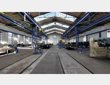 Ehemalige Fabrikhalle mit offenem Dach und Oberlichtern, geprägt von Stahlträgern und großen Fenstern an den Wänden. Die Halle enthält Werkbänke, Maschinen und Schienen entlang des Bodens für industrielle Anwendungen.