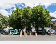 Offener Platz mit Kopfsteinpflaster, gesäumt von großen, dicht belaubten Bäumen. Im Vordergrund stehen Holztische, dahinter parken Foodtrucks und weitere Fahrzeuge; im Hintergrund sind städtische Gebäude sichtbar.