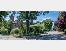 Straßenszene in einem Wohngebiet mit Kopfsteinpflaster, Bäumen und eingegrenzten Gärten. Im Hintergrund ist ein Backsteinturm mit spitzer Kuppel sichtbar, umgeben von niedrigen Häusern mit roten Ziegeldächern.