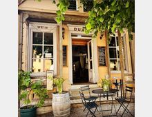 Gemütliches Bistro mit rustikaler Fassade, einschließlich bodentiefer Fenster und offener Tür, begleitet von handbeschriebenen Tafeln. Vor dem Eingang stehen ein Weinfass mit Blumen, kleine Metalltische und Stühle, sowie grüne Baumblätter, die Schatten spenden.