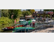 Hafenbereich mit kleinen Flussbooten, die teilweise mit grünen Planen abgedeckt sind, neben einer Promenade mit einem Restaurant mit Terrasse im Hintergrund. Im Vordergrund befindet sich eine gepflasterte Rampe mit Geländern und im Hintergrund ein kleiner Wasserfall sowie Bäume.
