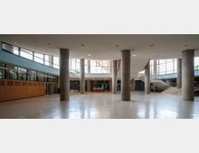 Großer, moderner Innenraum mit hohen Decken, Betonstützen und viel Tageslicht, das durch große Fensterfronten einfällt. Eine breite Treppe mit Geländern führt auf eine Empore, während der Boden hell und glatt gestaltet ist.