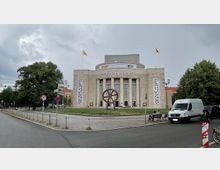 Die Aufnahme zeigt die Volksbühne in Berlin, ein großes klassizistisches Theatergebäude mit Säulenfassade und seitlichen Bannern. Im Vordergrund steht eine moderne Skulptur aus Metall in Form eines großen Rads, umgeben von einer Grünfläche und Gehwegen.