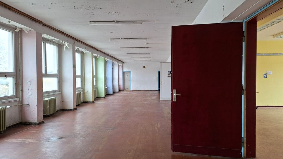 Langer, leerer Flur in einem älteren Gebäude mit großen Fenstern und Heizkörpern entlang der linken Wand sowie abblätternder Farbe an Decke und Wänden. Rechts ist eine geöffnete dunkelrote Tür zu einem angrenzenden Raum zu sehen.