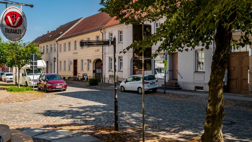 Blick auf eine gepflasterte Innenstadtstraße mit geparkten Autos, einem Straßenschild „Dammstraße“ und einer runden „Tierarzt“-Leuchtreklame am linken Gebäude. Im Hintergrund stehen zweigeschossige, helle Häuser mit roten Ziegeldächern und Café-Fassade, begleitet von Straßenbäumen am Rand.