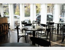 Innenraum eines modernen Restaurants mit dunklen Holztischen und schwarzen Stühlen auf Holzboden. Große Fenster mit weißen Vorhängen lassen Tageslicht herein und geben den Blick auf parkende Autos und Bäume außerhalb frei.