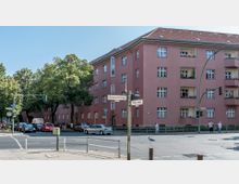 Wohnhaus mit vier Stockwerken in rosa Außenfassade an einer Straßenecke, umgeben von Bäumen und einer asphaltierten Straße mit Verkehrsampeln. Im Vordergrund Straßenschilder mit den Namen „Birkbuschstraße“ und „Klingsorstraße“ sowie parkende Autos und einige Passanten.
