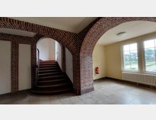 Treppenhaus mit roten Backsteinbögen, einem dunkelbraunen Holztreppenaufgang und großen Fenstern, die auf eine grüne Außenlandschaft blicken. Der Boden ist mit hellen Fliesen ausgelegt, und an der Wand hängt ein Feuerlöscher.