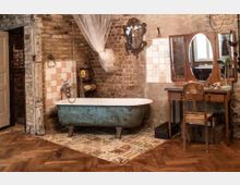 Rustikales Badezimmer mit hellblauer freistehender Badewanne auf dekorativen Fliesen, umgeben von freiliegenden Backsteinwänden. Daneben steht ein antischer Frisiertisch mit Spiegeln und einem hölzernen Stuhl auf einem Fischgrät-Holzboden.