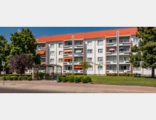 Mehrfamilienhaus mit vier Stockwerken, weißer Fassade und roten Dachziegeln, mit Balkonen, von denen einige mit Markisen und Sonnenschirmen ausgestattet sind. Im Vordergrund befinden sich eine Bushaltestelle mit überdachtem Wartebereich, Gehwege, Grünflächen und mehrere Bäume.