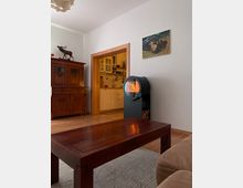 Wohnzimmer mit Holzboden, großem dunklen Couchtisch und einem modernen schwarzen Kaminofen mit Sichtfenster; daneben steht ein antiker Holzschrank. Durch einen breiten Türrahmen ist eine helle Küche mit weißen Schränken und Fliesenspiegel zu sehen, an der Wand hängt ein Gemälde.